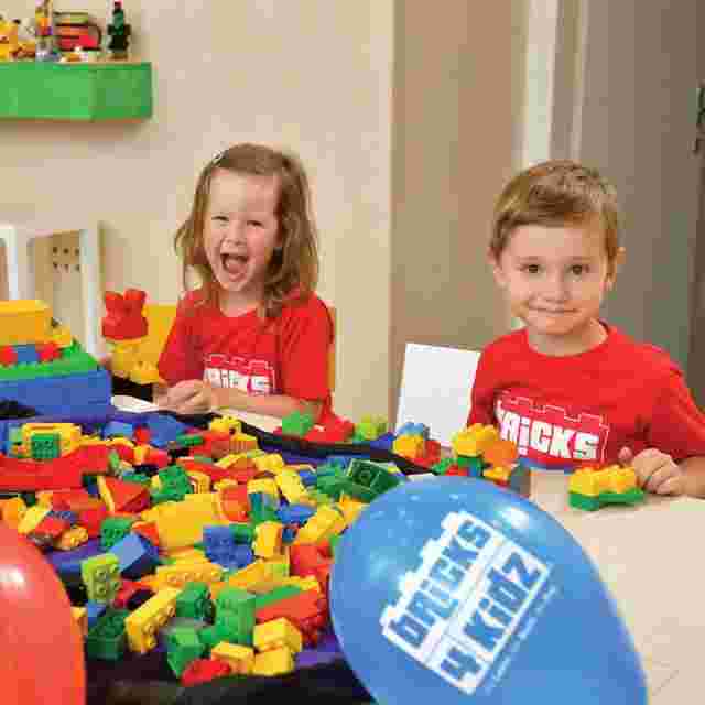 Two children in red shirts play with colorful building blocks at a table. A blue balloon with "bricks 4 kidz" text is visible in the foreground.
