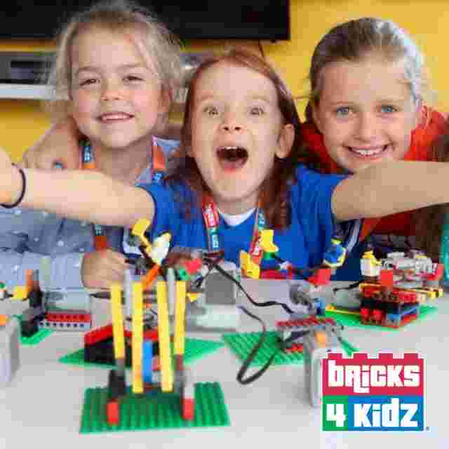 Three children excitedly play with colorful building blocks at a table. The logo "Bricks 4 Kidz" is in the bottom right corner.