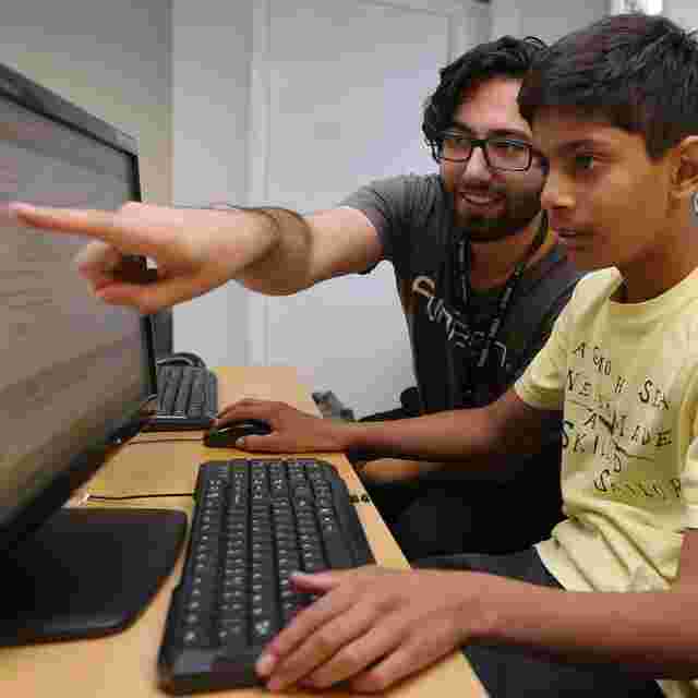 A man guides a boy on a computer, pointing at the screen. The boy is focused, sitting at a desk with a keyboard and monitor.