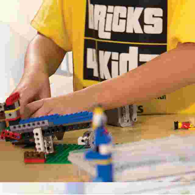 Child building a LEGO structure on a table, wearing a yellow "Bricks 4 Kidz" shirt.