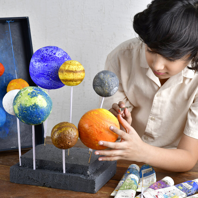 A child carefully paints a handmade model of the solar system, with colorful planets mounted on sticks, surrounded by paint tubes on a wooden table.
