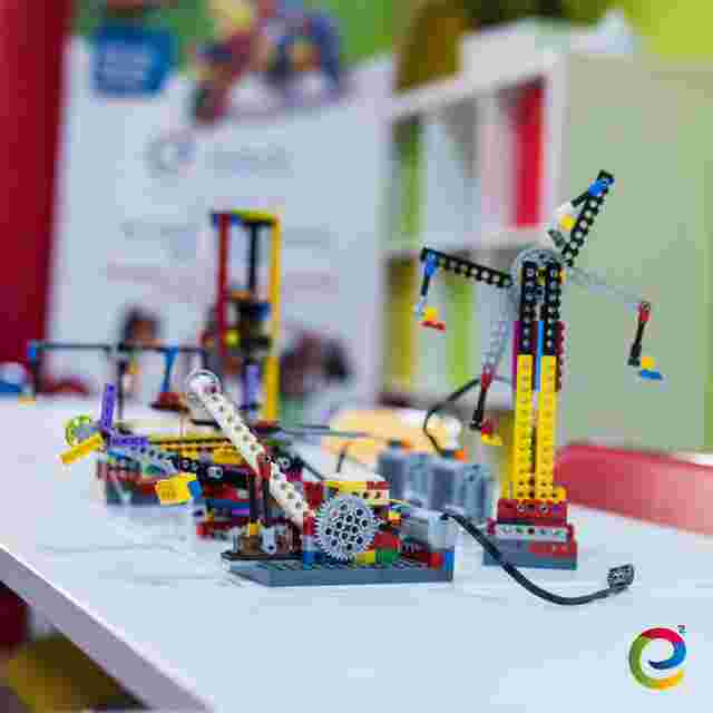 Colorful LEGO robotics models on a white table, with a blurred background of educational materials and a logo in the corner.