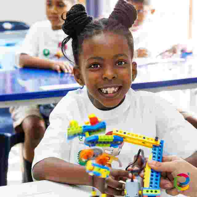 A smiling child with braided hair plays with colorful building blocks at a table, surrounded by other children in a classroom setting.