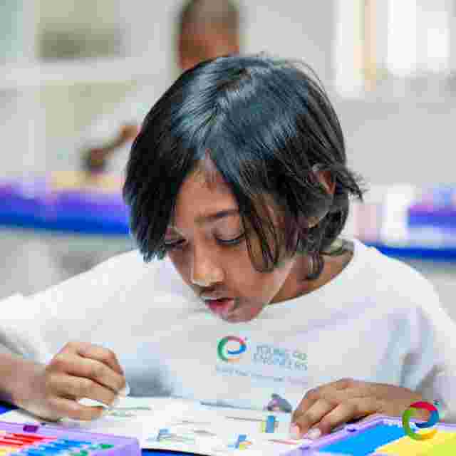 Child focused on assembling a project at a table, wearing a "Young Engineers" shirt, with colorful educational materials around.