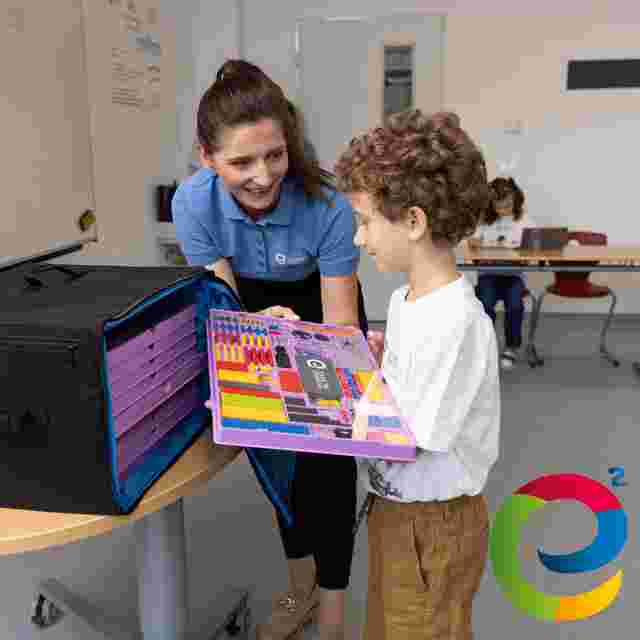 A teacher hands a colorful educational kit to a young boy in a classroom. Other students are seated at a table in the background.