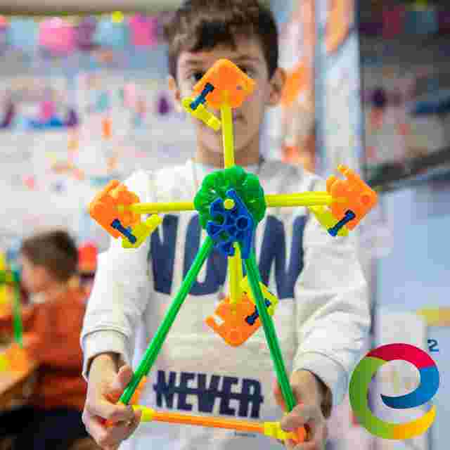 Child holding a colorful geometric structure made from plastic connectors in a classroom setting, with another child blurred in the background.