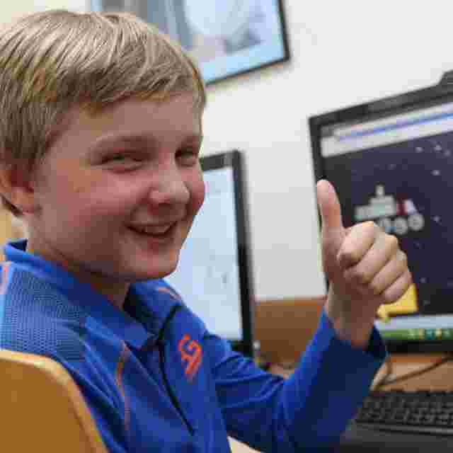 Smiling boy in a blue jacket giving a thumbs-up while sitting at a computer desk with screens displaying graphics.