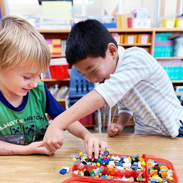 Two boys happily playing with colorful building blocks at a classroom table, surrounded by shelves of books and educational materials.
