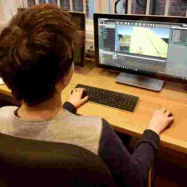 A person with short brown hair is sitting at a desk, using game design software on a computer with a large monitor.