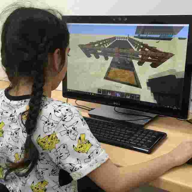 Girl with braided hair plays Minecraft on a desktop computer, wearing a Pikachu-patterned shirt.