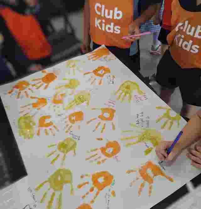 Children wearing "Club Kids" vests paint handprints on paper, writing names and messages in colorful inks.