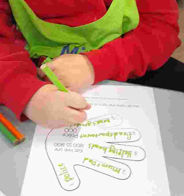 A child in a red sweater writing on a worksheet with a hand outline, using a green marker at a table.
