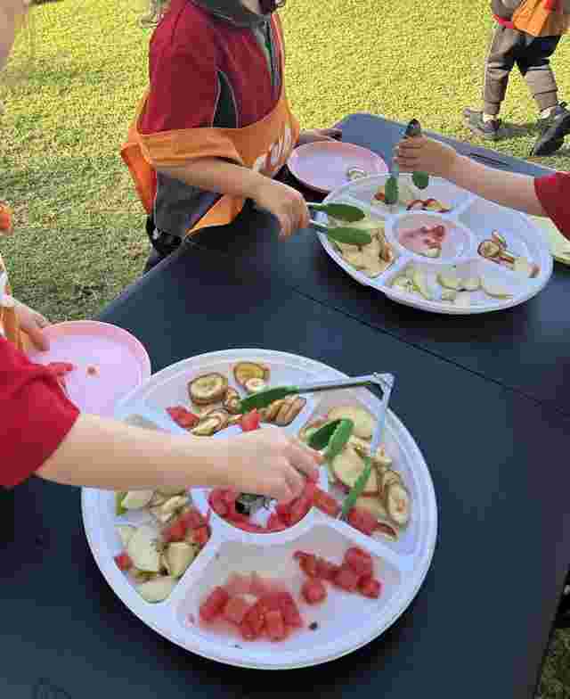 Children serving themselves fruit slices and watermelon chunks from platters using tongs at an outdoor setting.