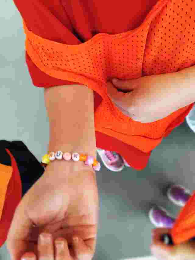 A child wearing an orange sports bib shows a beaded bracelet with the letters "RUKN" on their wrist.