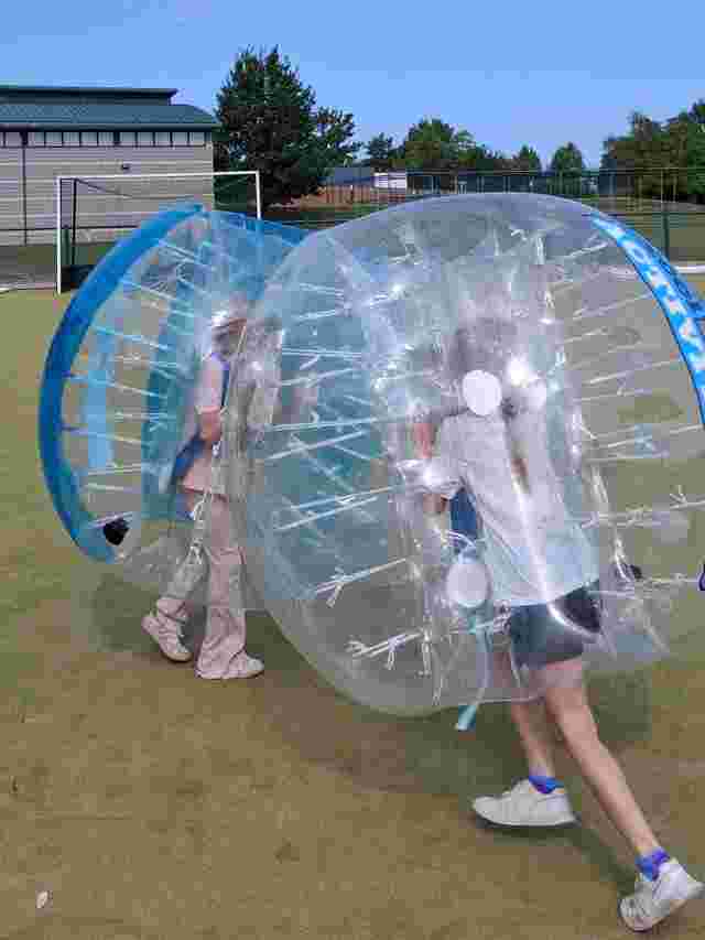 Two people in inflatable bumper balls on a grassy field, with a building and trees in the background under a clear blue sky.