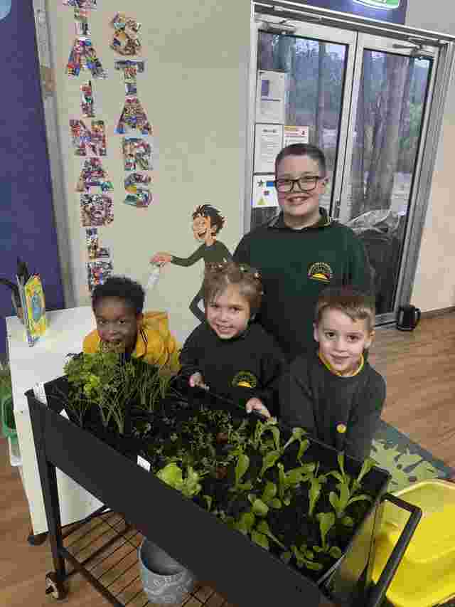 Four children smiling around a small indoor garden with various plants, in a classroom setting.