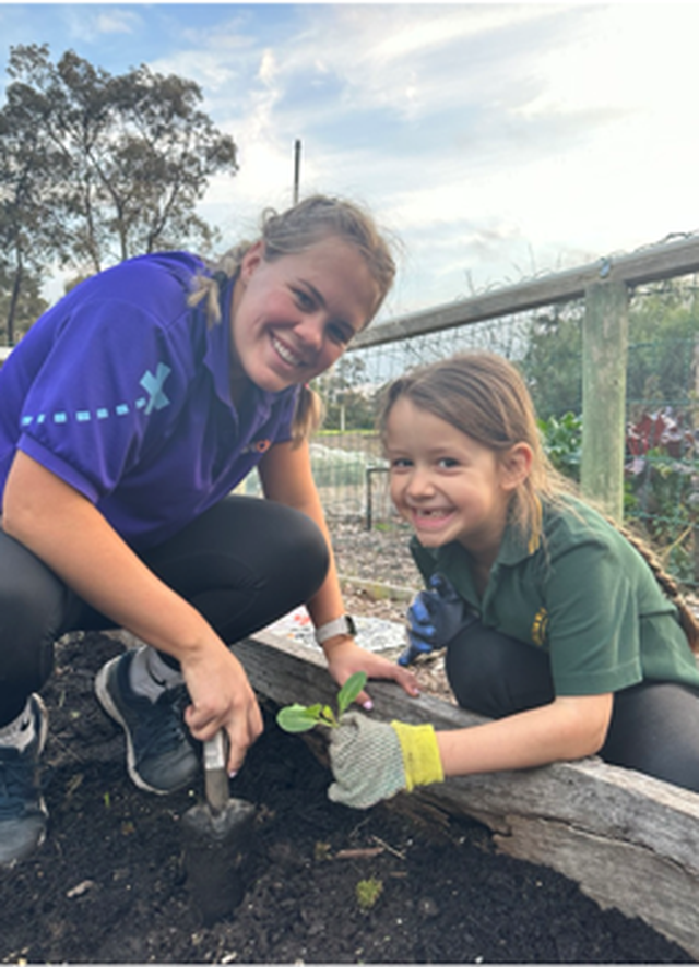 Two people gardening together, smiling while planting in soil, with one holding a trowel and the other wearing gloves. Trees and fencing are in the background.
