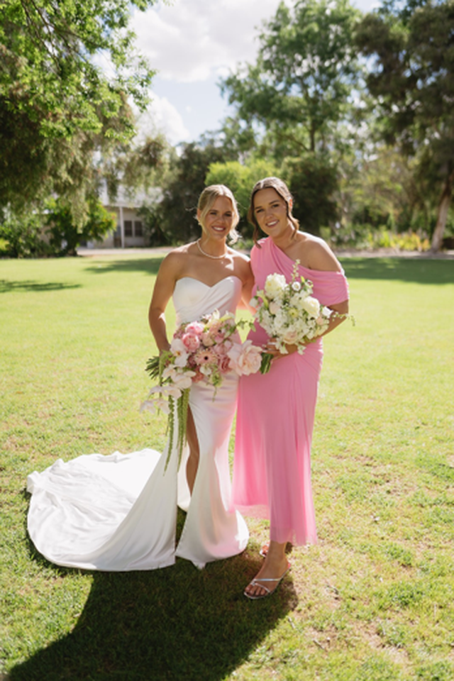 Two women smiling outdoors, one in a white wedding dress with flowers, the other in a pink dress, both holding bouquets, standing on grass.
