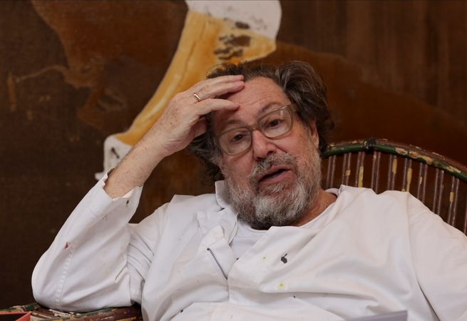 Julian Schnabel touching his forehead while sitting in a chair, talking to the camera