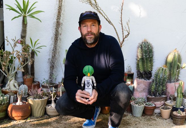 man in baseball cap crouching down holding a cactus man action figure
