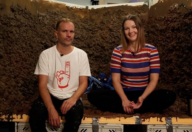 two artists sitting inside an installation made out of soil and looking into the camera