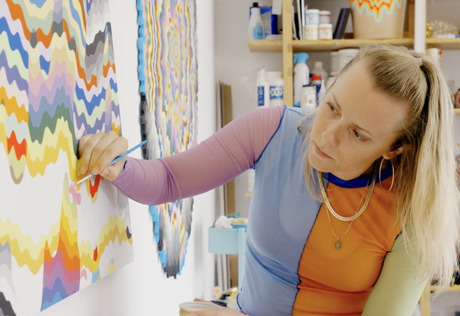 artist wearing colourful long-sleeved top concentrating on an unfinished painting in front of her which she holds a small paintbrush to