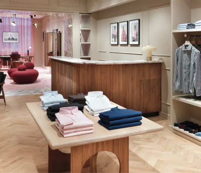 The inside of a Thomas Pink store. There are collared button-up shirts on a wooden table and others hanging on the rack. Red round chairs are in the back of the store near the dressing rooms.