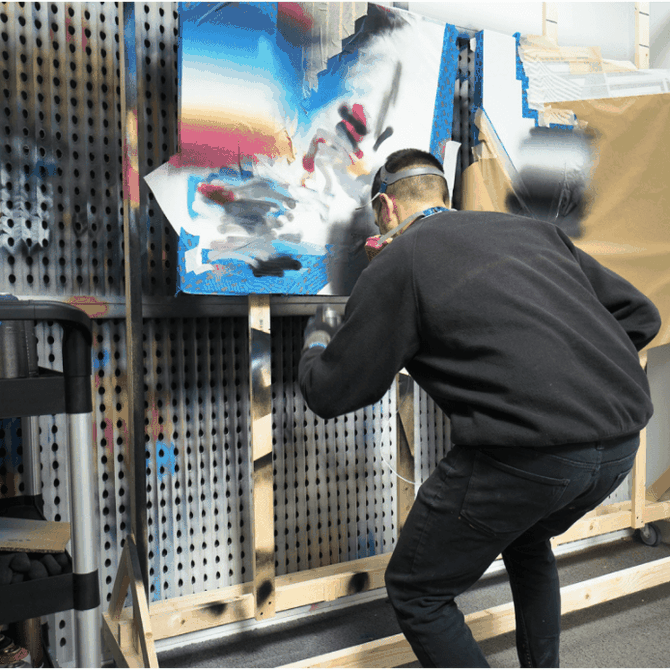 man with mask on as he adds paint to a colourful canvas hung up in front of him