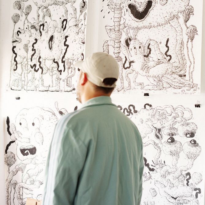 artist with his back turned to the camera looking towards four large drawings on paper which are pinned to the wall of his studio