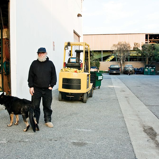Paul McCarthy in black hoodie and navy cap, standing with a dog outside an industrial warehouse building