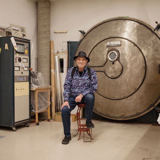 Larry Bell sat on a stool smiling in his glass work studio