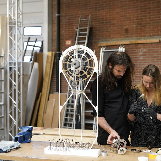 male and female artist inspecting an object in the female artists hands, amongst electrical circuits and contraptions in their studio