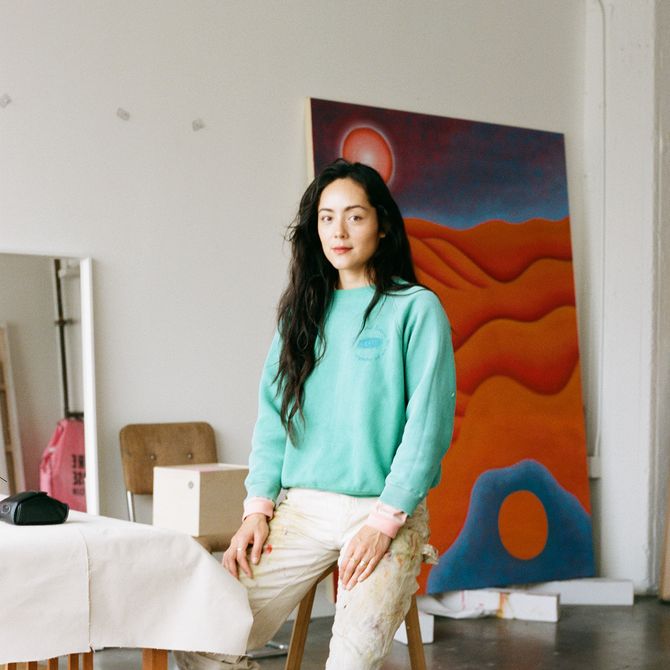 Camilla Engström sitting on a stool in her studio, blue and orange painting in the background
