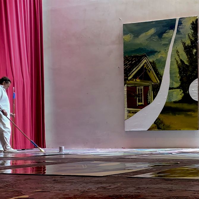 artist wearing white overalls, using a long brush to paint a print on the floor of his large studio space – enclosed on the left by a scarlet curtain