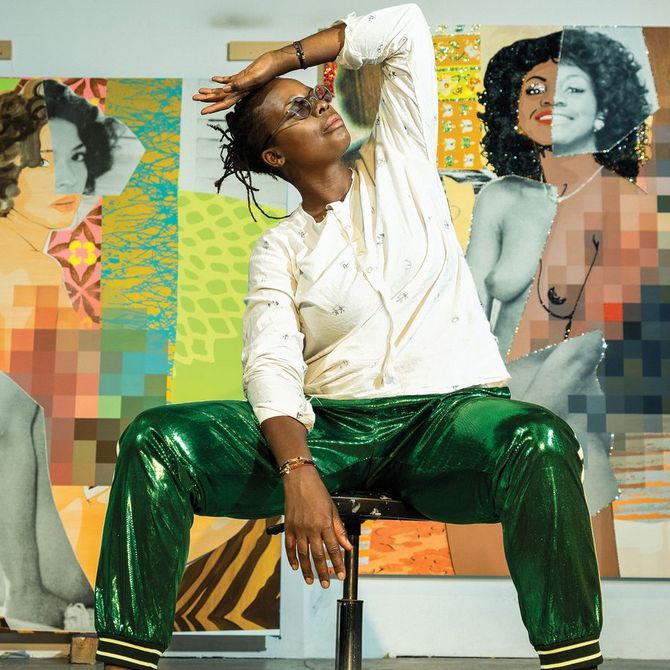 Mickalene Thomas sat on a stool in front of two paintings raising her hand to her head