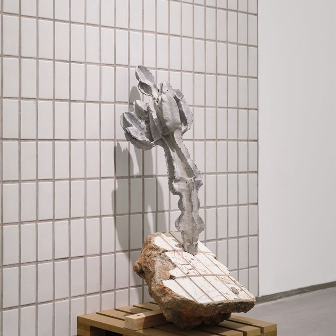 installation view of tiled grid wall with plant sculpture leaning against it balanced on a concrete block