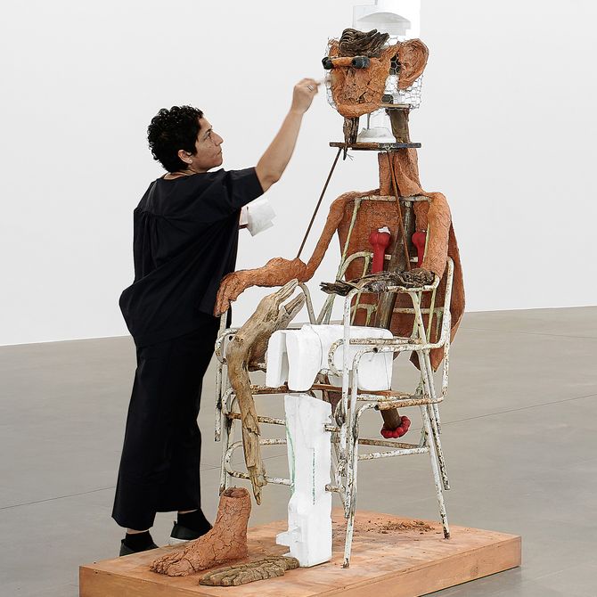Huma Bhabha reaching upward to adjust the head of a sculpture