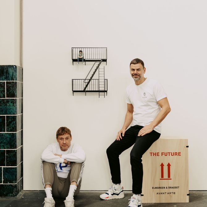 Elmgreen & Dragset sat on the floor and on a box in their studio next to a small sculpture hung on the wall