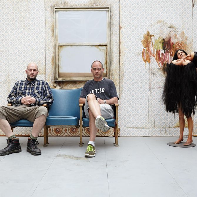 Jake and Dinos Chapman sat side-by-side in a room, empty other than a four headed sculpture