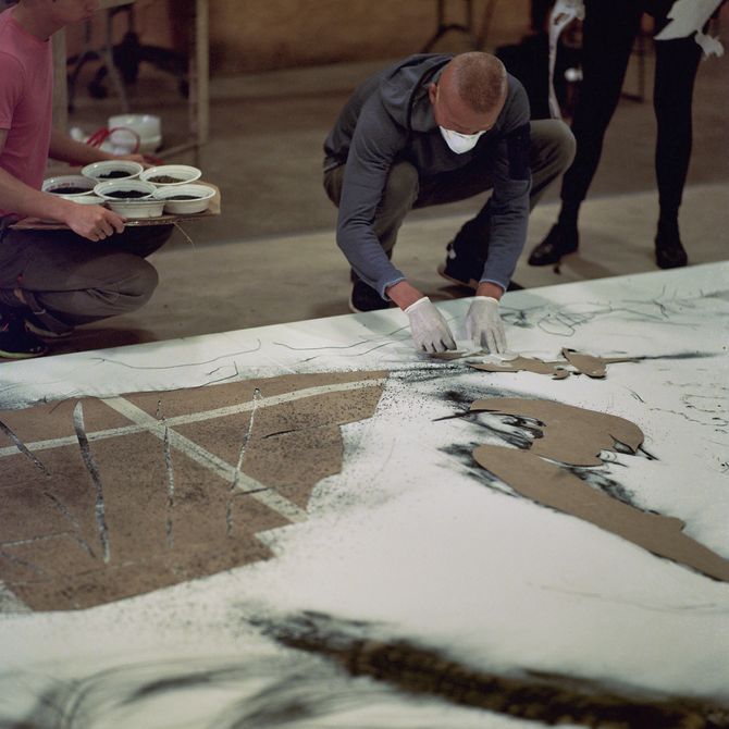 artist crouching down wearing a white medical mask and white latex gloves whilst he touches the white painted wood before him and an assistant kneels at his side with bowls of paint