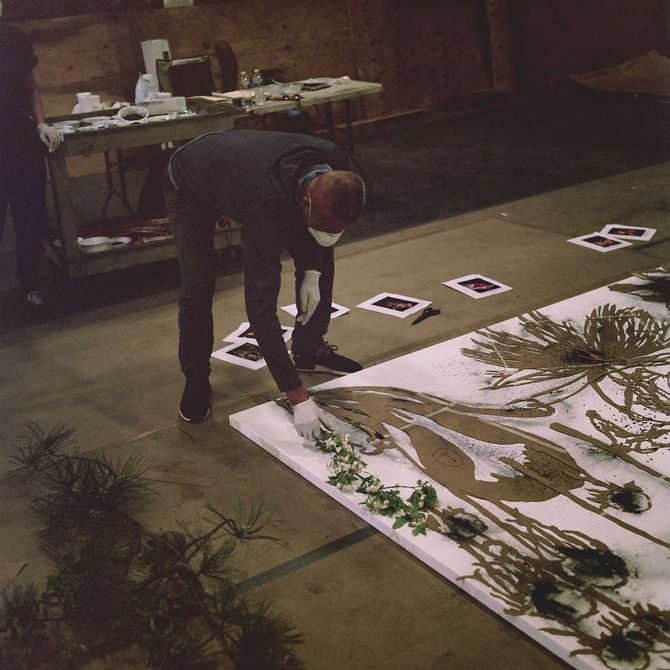 artist bending down to hold a flower up to a large wooden canvas with outlines of flowers and plants on it