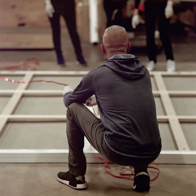 artist with back facing camera as he kneels down to the floor and looks towards several assistants in front of him, whilst holding a long piece of red wire