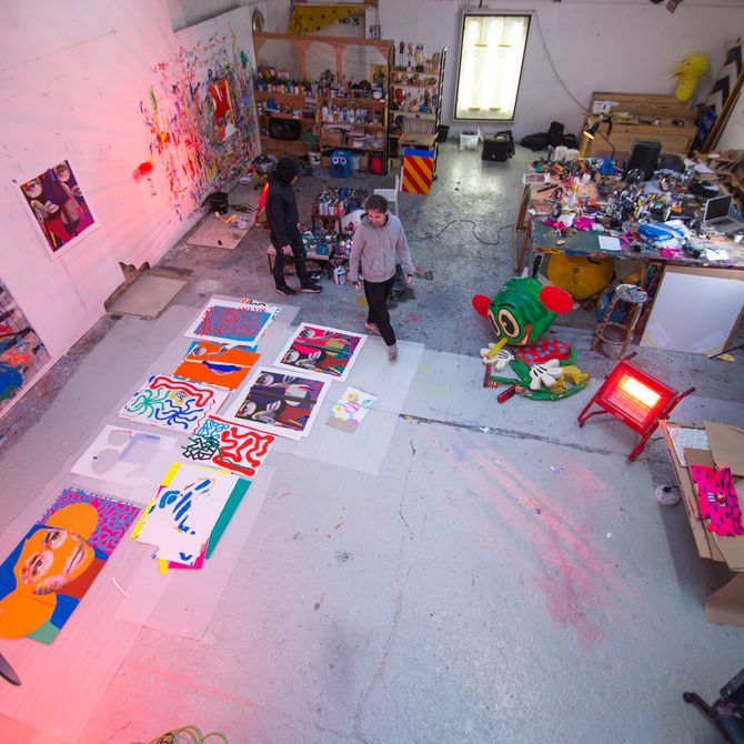 view looking down on the artists studio, in which the artist walks around a collection of paintings and prints laid out on the floor to his right