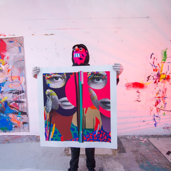 artist wearing a pink mask covering his face with a black hood over the top, as he stands in his studio holding up a large print of two collage faces and bright blocks of colour