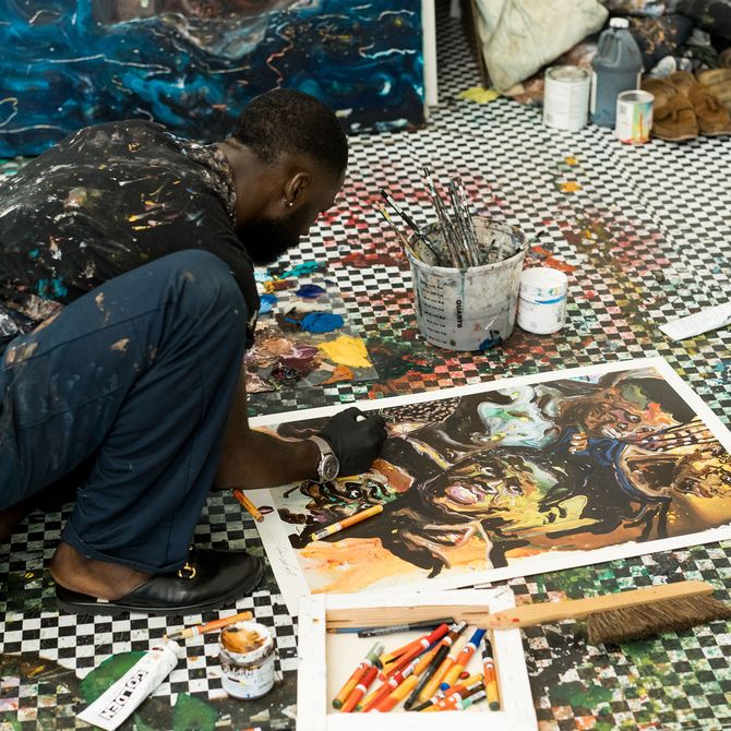 Ludovic Nkoth crouching on the floor surrounded by paints and pens as he adds details to a painting on the floor