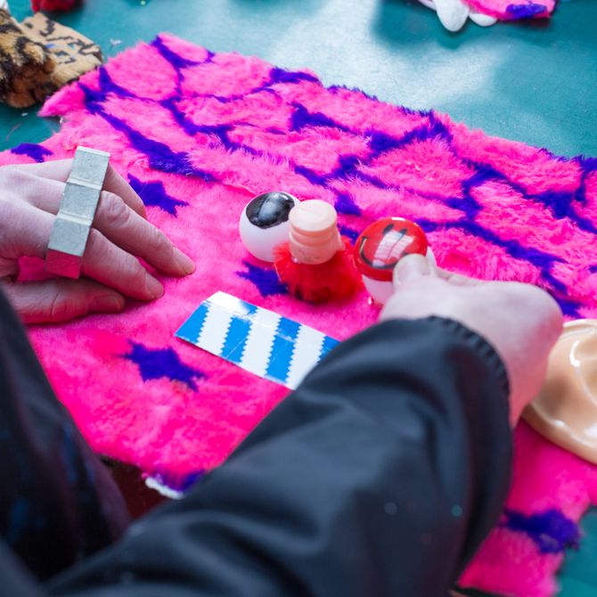 Close-up of artists hands as he works on a pink piece of cloth which he decorates with blue patterns, sticking on an ear and an eye and a red button