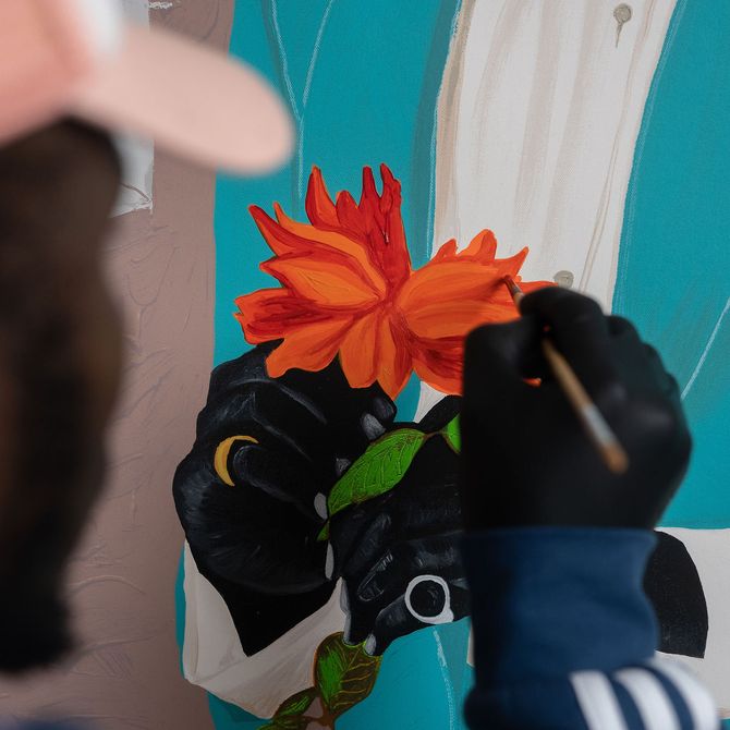 artist hand holding a paintbrush up to a painting to add detail to an orange flower