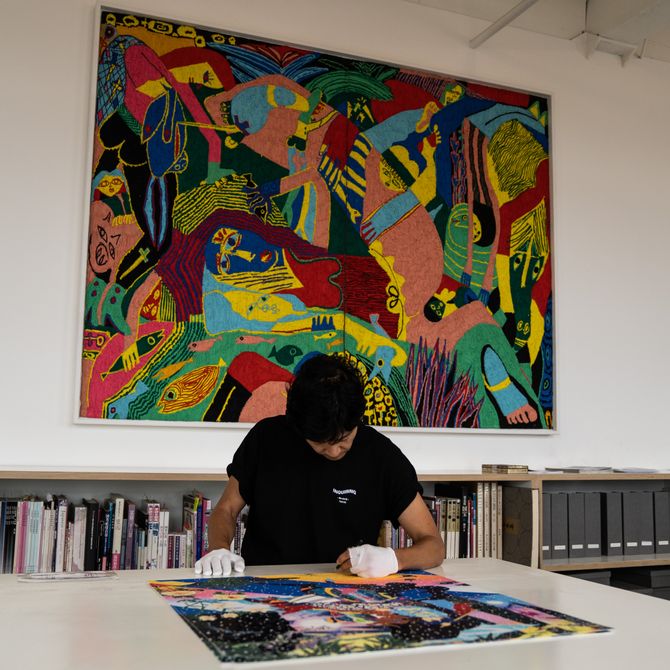 artist in studio looking down at artwork on table, with a large scale painting and bookshelves behind him