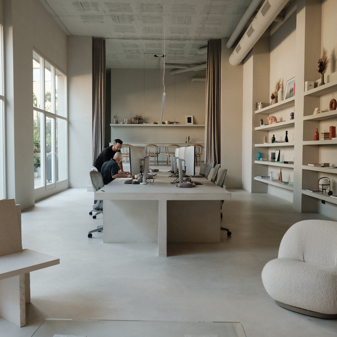 Cream coloured studio with large desk in the centre, and bookshelves on the right wall. Two seats in the foreground