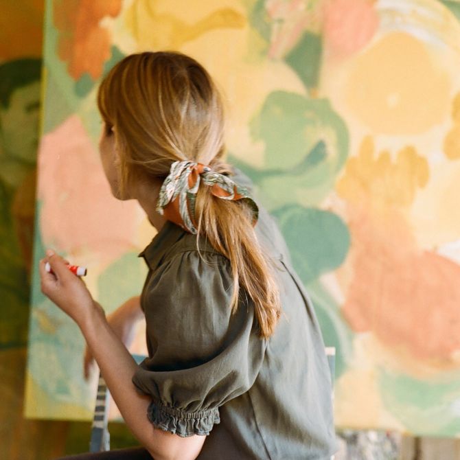 artist with back turned to camera whilst casts gaze upon a pastel coloured painting with a ribbon tying her hair back